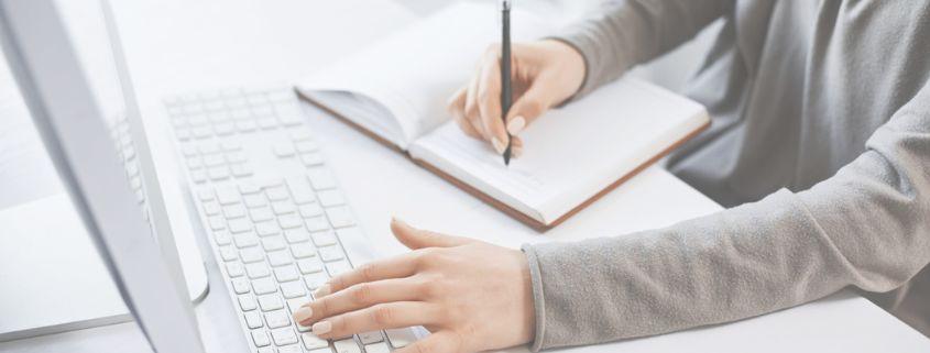cos-e-il-copywriting-e-perche-e-importante Una persona è intenta a scrivere in un quaderno, seduta a un tavolo con il suo pc portatile davanti